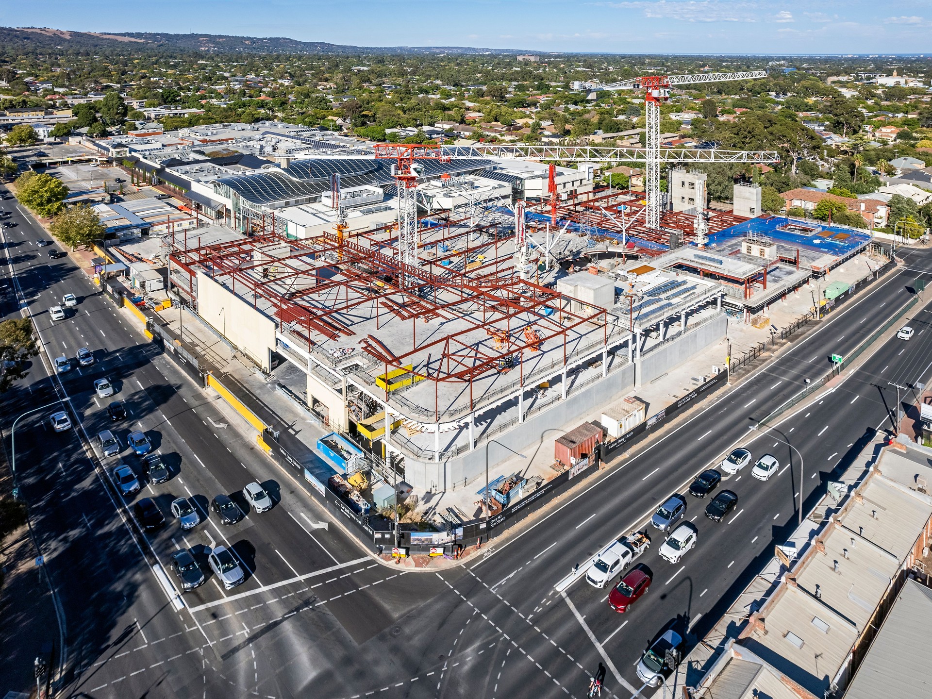 Aerial view retail shopping mall construction site, two tower cranes & equipment in Adelaide's eastern suburbs, busy main road intersection with traffic buildup on public holiday.